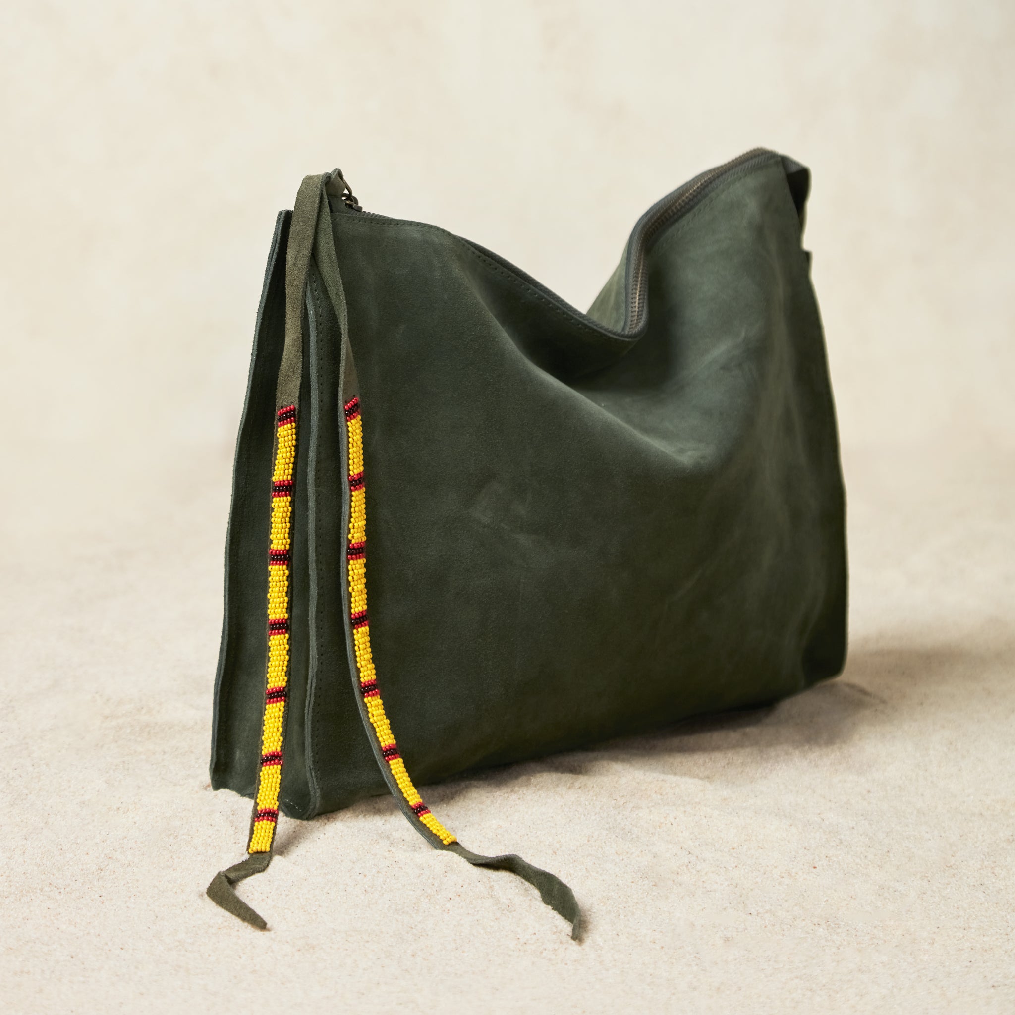 Alternative diagonal view of large olive green suede leather pouch bag with yellow, black, and red beaded zipper pull ties on a sand background