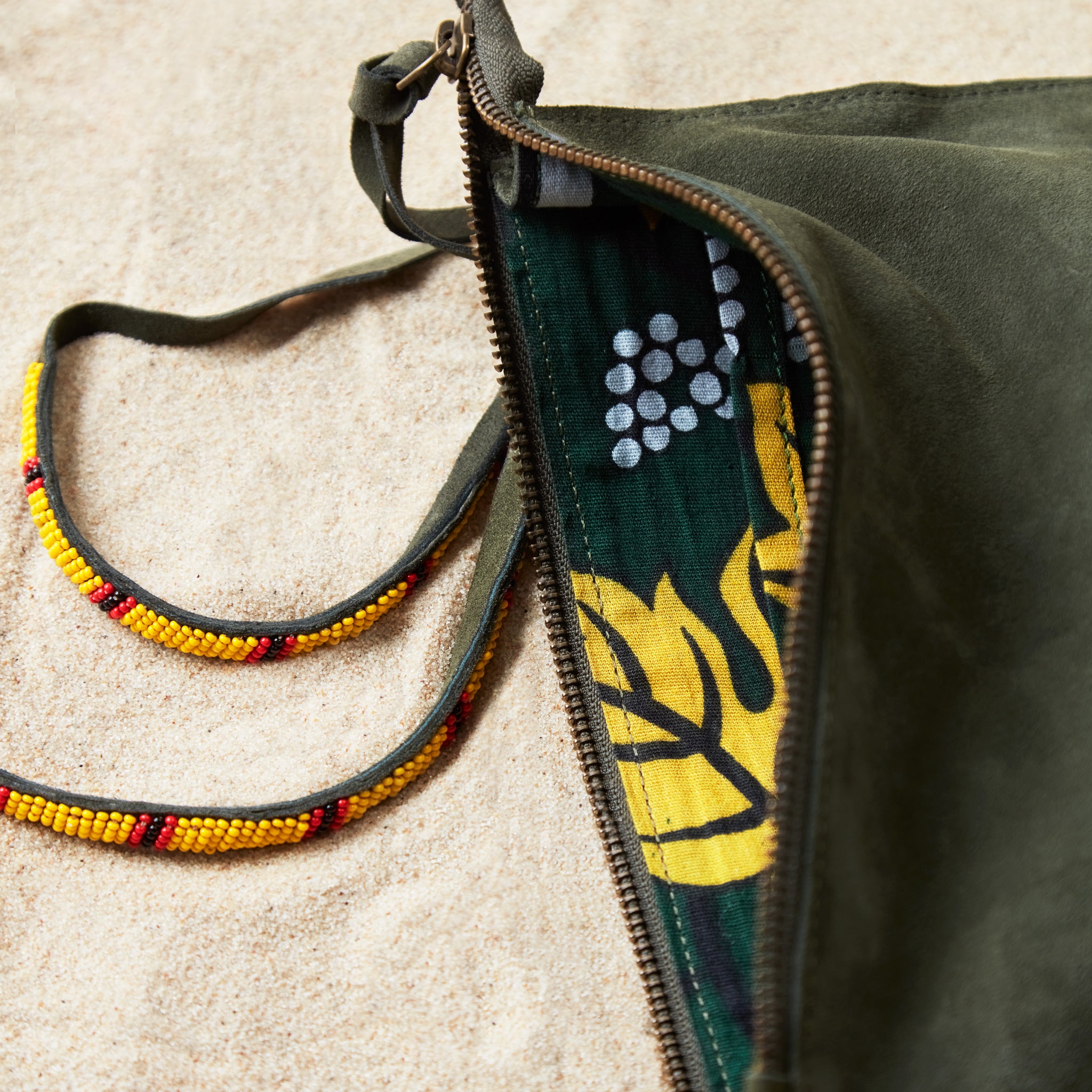 Close up view of colorful kanga fabric lining of small olive green suede leather pouch bag with yellow, black, and red beaded zipper pull ties on a sand background