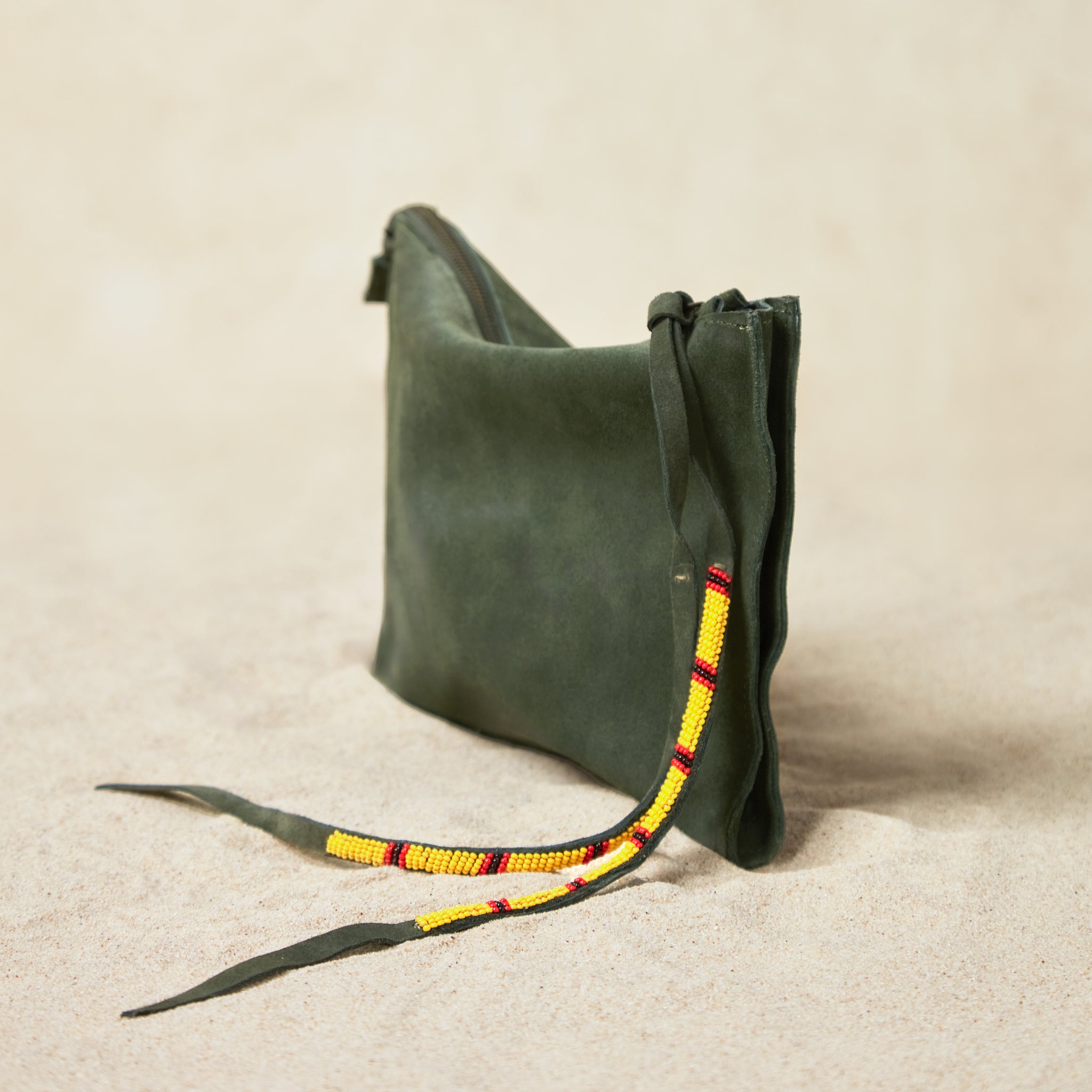 Alternative diagonal view of small olive green suede leather pouch bag with yellow, black, and red beaded zipper pull ties on a sand background