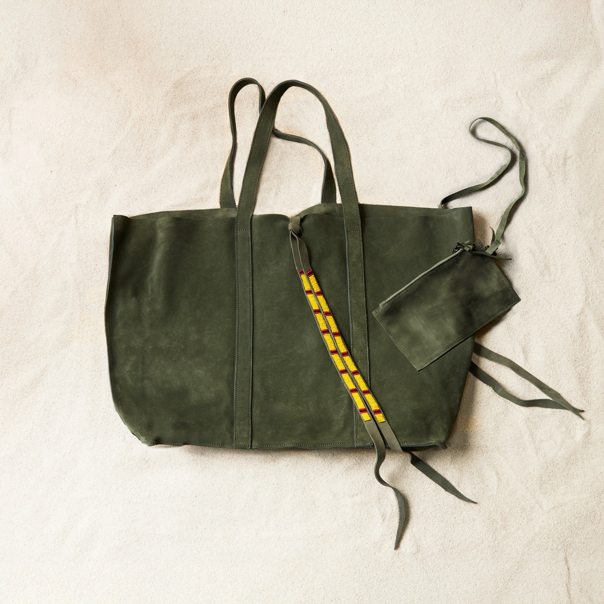 Flat view of olive green suede leather tote bag with yellow, black, and red beaded tassel ties including an attached interior zipper pouch on a sand background