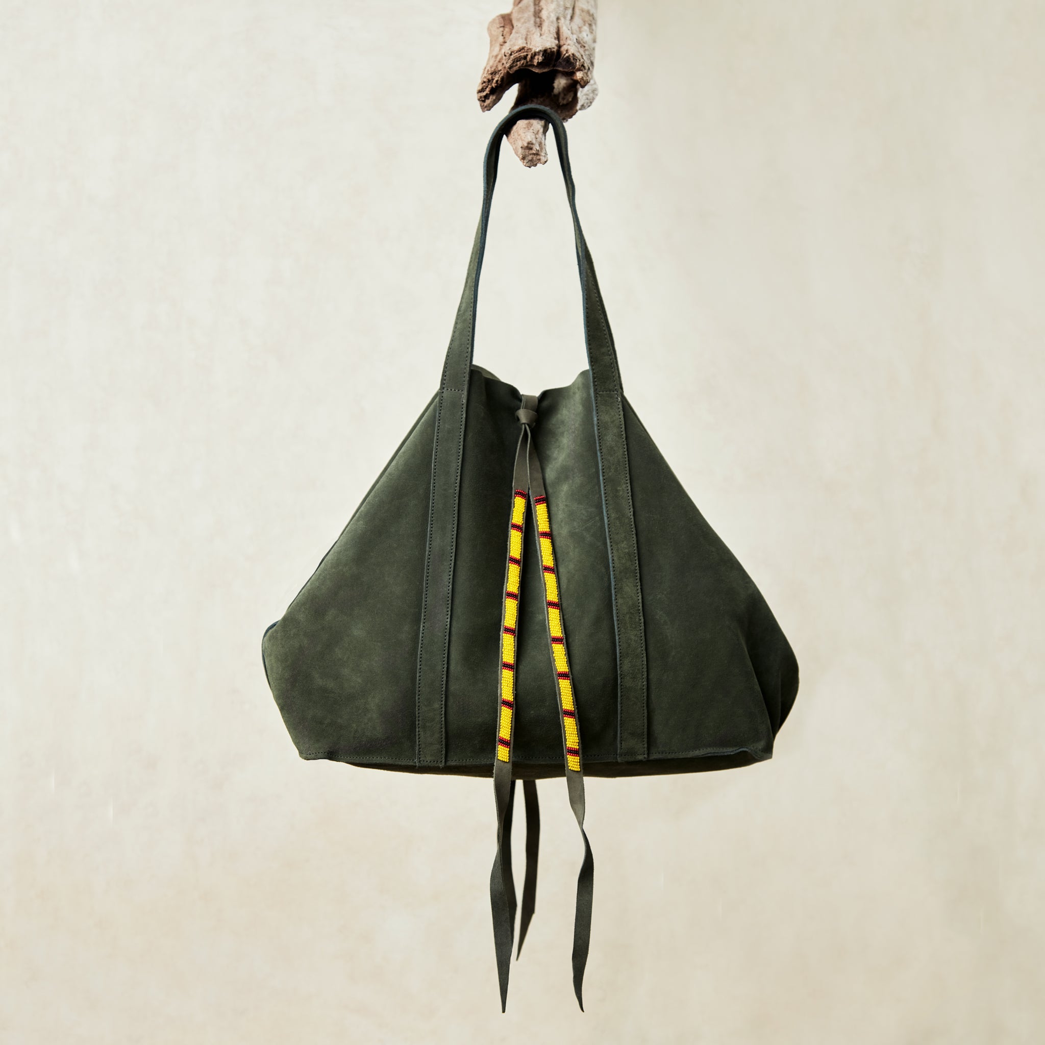 Olive green suede leather tote bag with yellow, black, and red beaded tassel ties hanging on a branch with a beige background