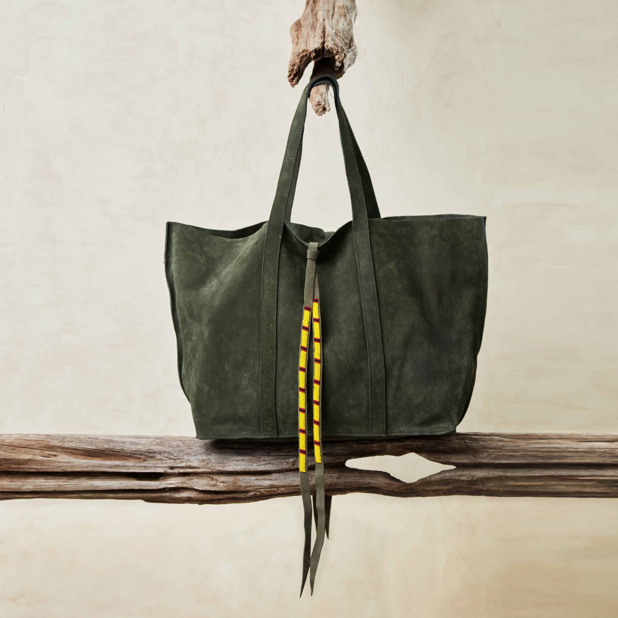 Olive green suede leather tote bag with yellow, black, and red beaded tassel ties hanging and sitting on wooden branches with a sand background