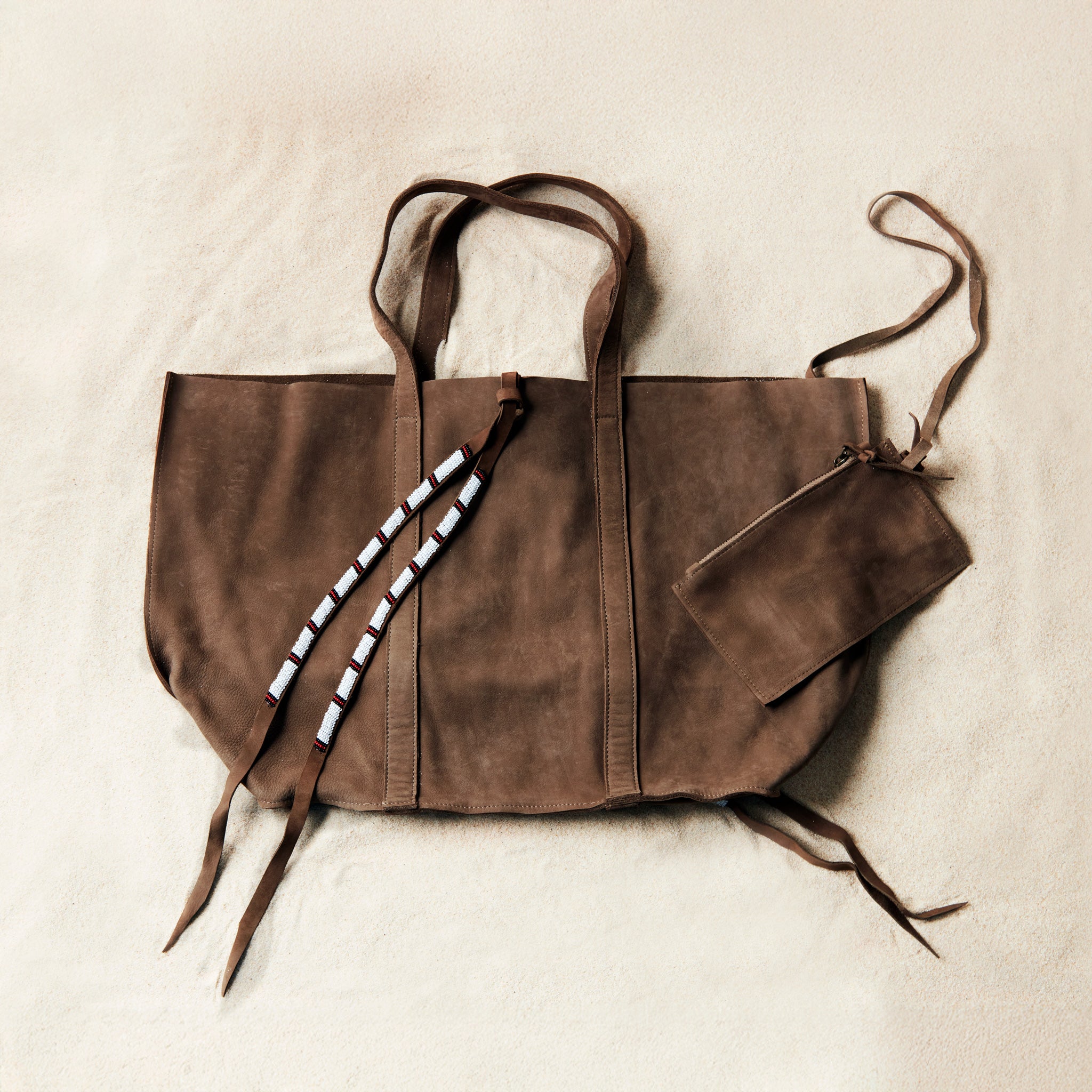 Flat view of brown sand suede leather tote bag with white, black, and red beaded tassel ties including an attached interior zipper pouch on a sand background