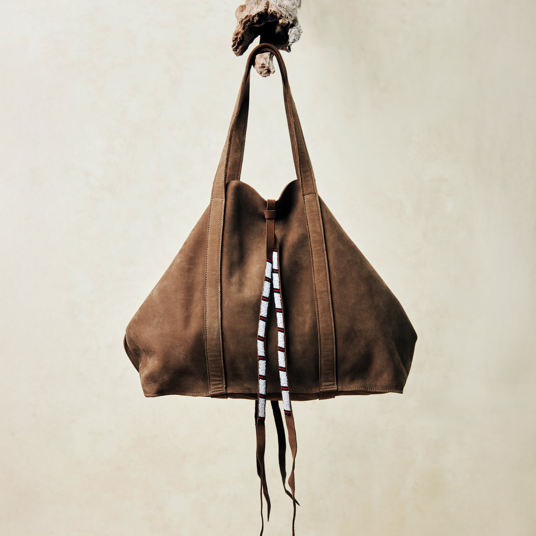 Brown sand suede leather tote bag with white, black, and red beaded tassel ties handing on a branch with a beige background