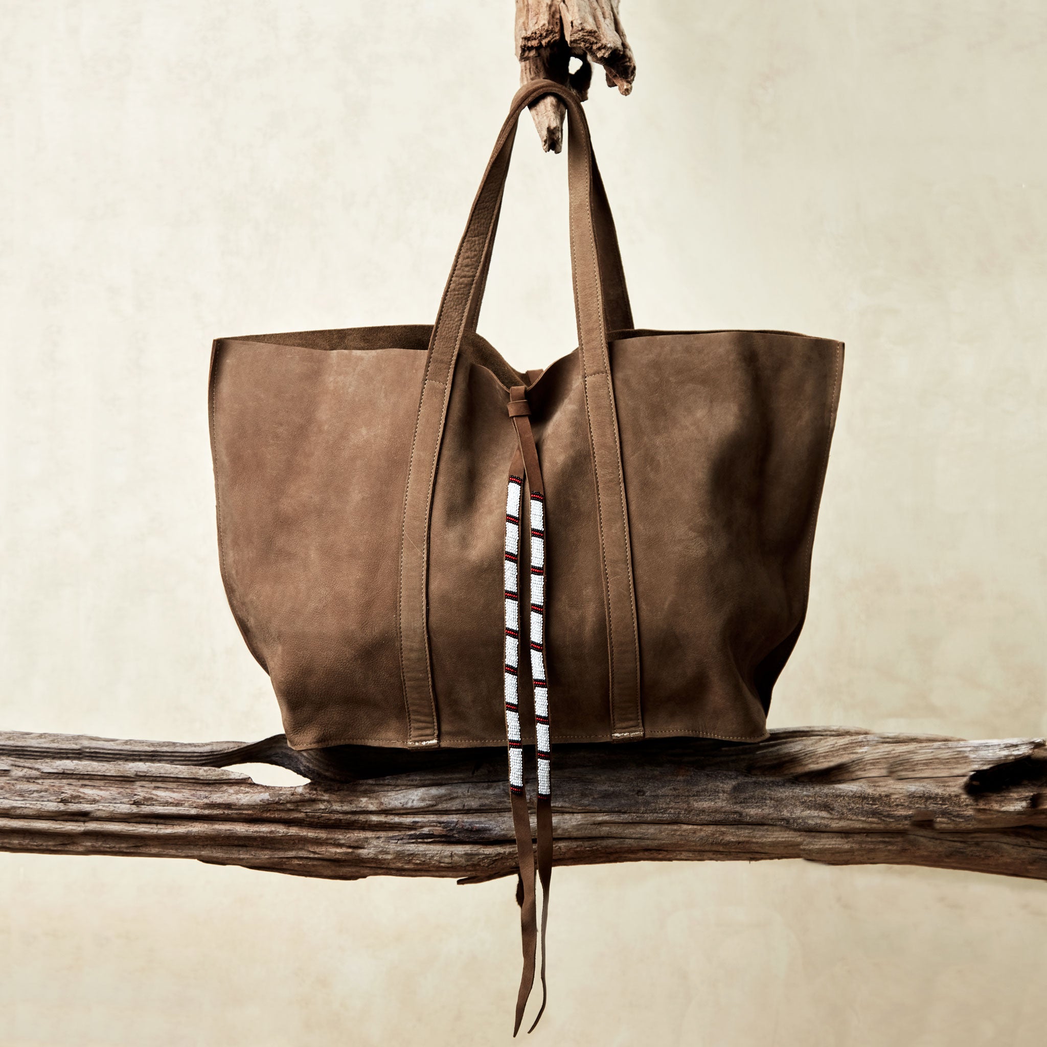 Brown sand suede leather tote bag with white, black, and red beaded tassel ties on a wooden branch with a beige background