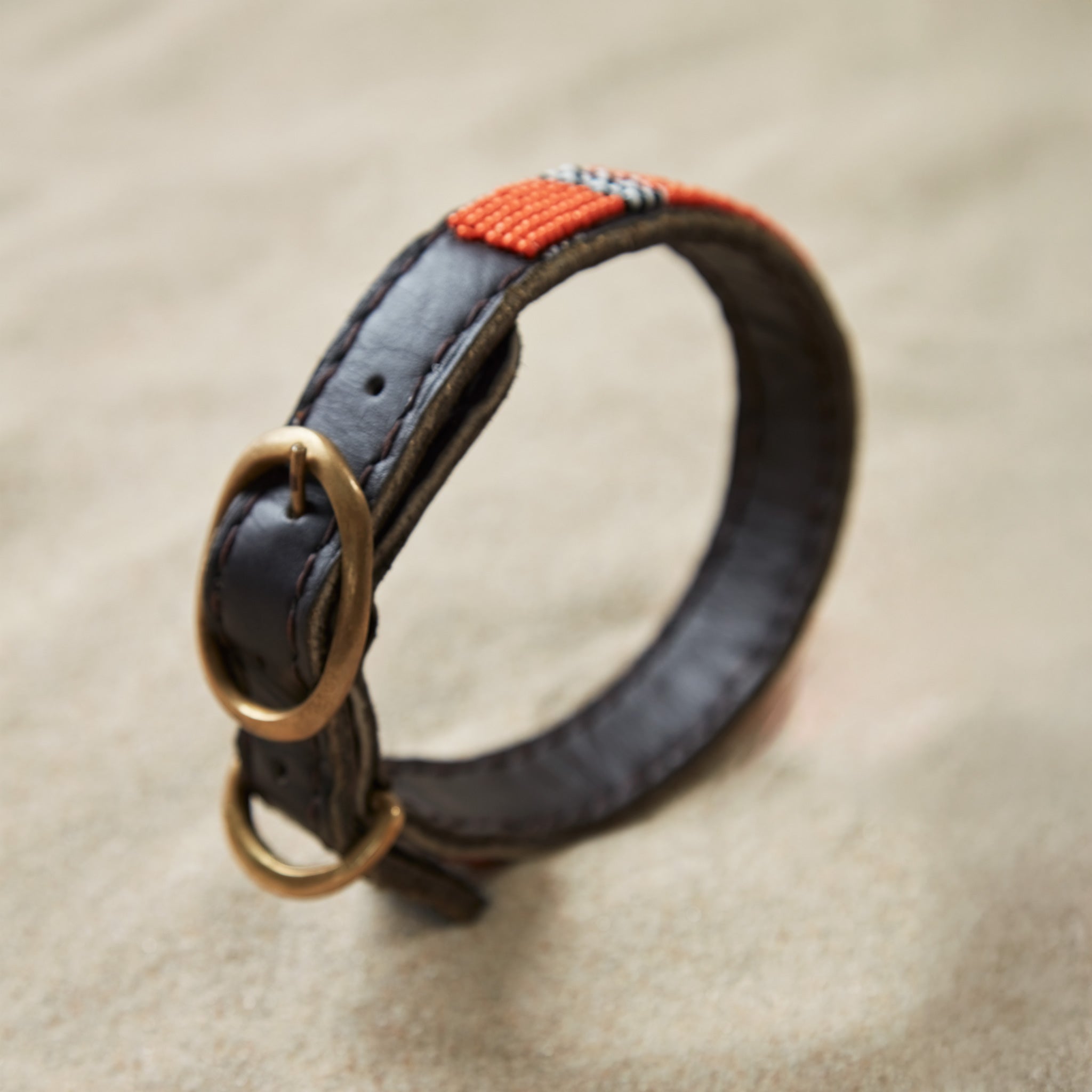 Alternative view of a brown leather dog collar with orange, black, and white beads and a brass buckle on a sand background