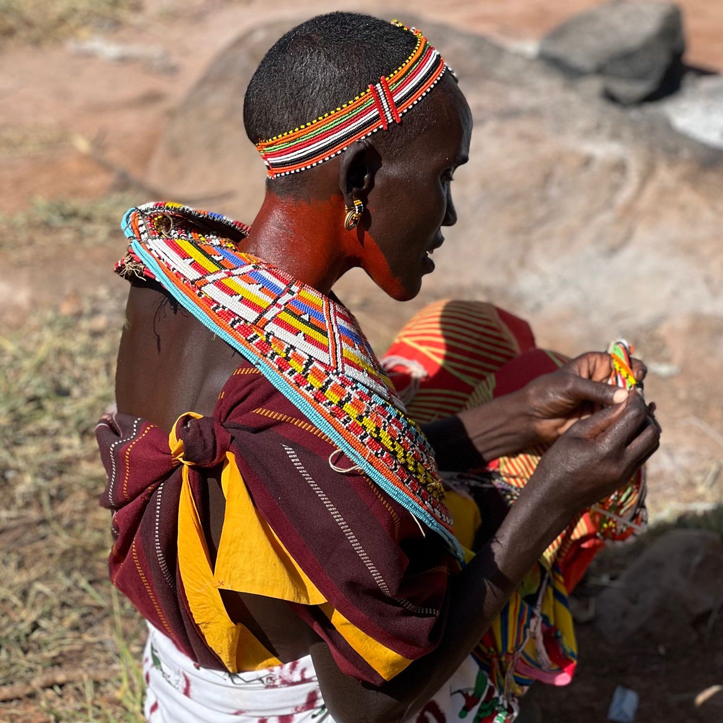 Woman in colorful traditional attire with a large beaded neck piece and headband working on a project sitting on the ground with rocks in the background