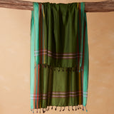 An olive green cotton sarong/pareol with colorful striped borders and tassels, hanging from a rustic wooden beam against a warm beige background.