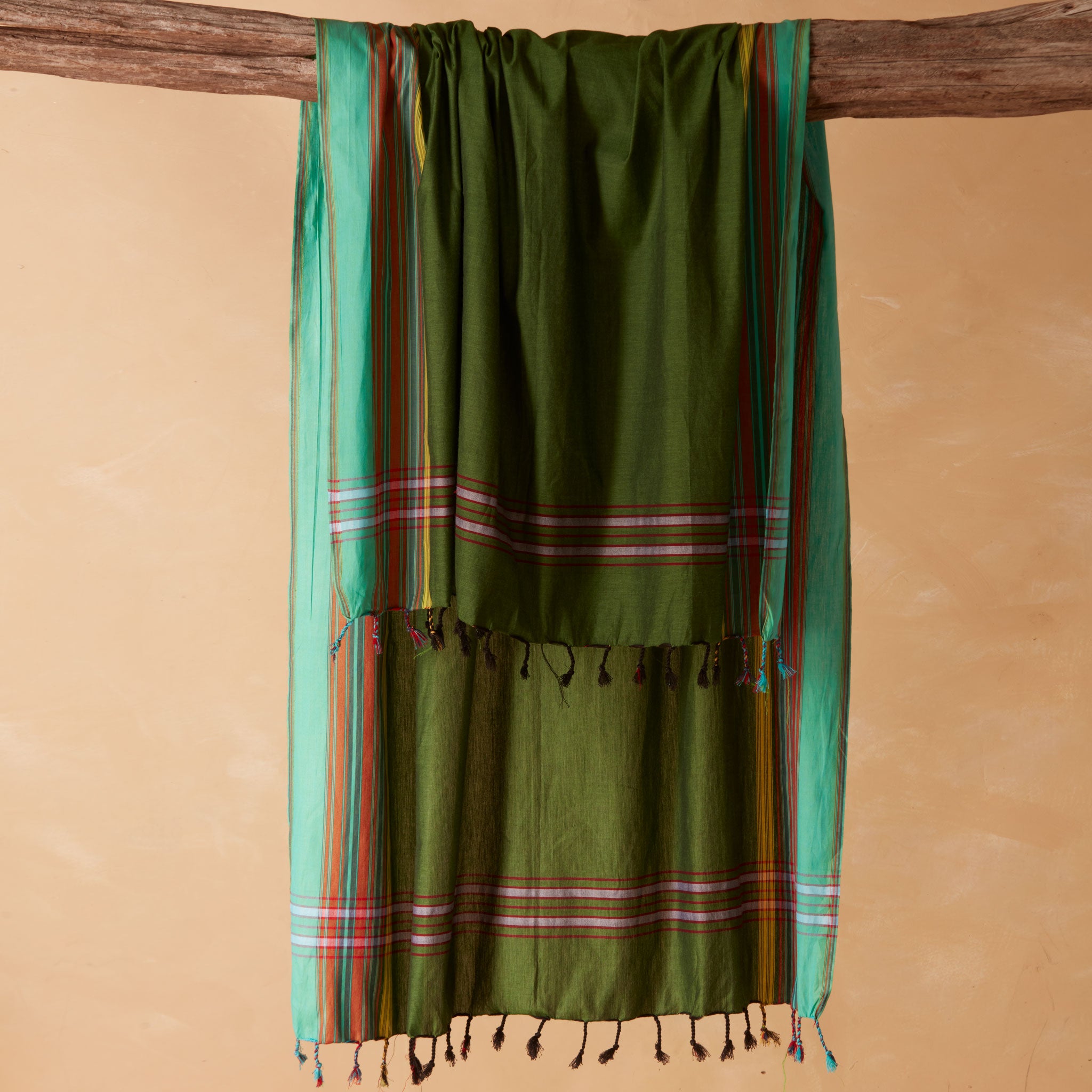 An olive green cotton sarong/pareol with colorful striped borders and tassels, hanging from a rustic wooden beam against a warm beige background.