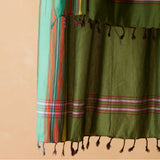 A close-up of an olive green sarong/pareo with horizontal striped borders in red, white, and pink, finished with dark tassels, shown against a warm beige background.