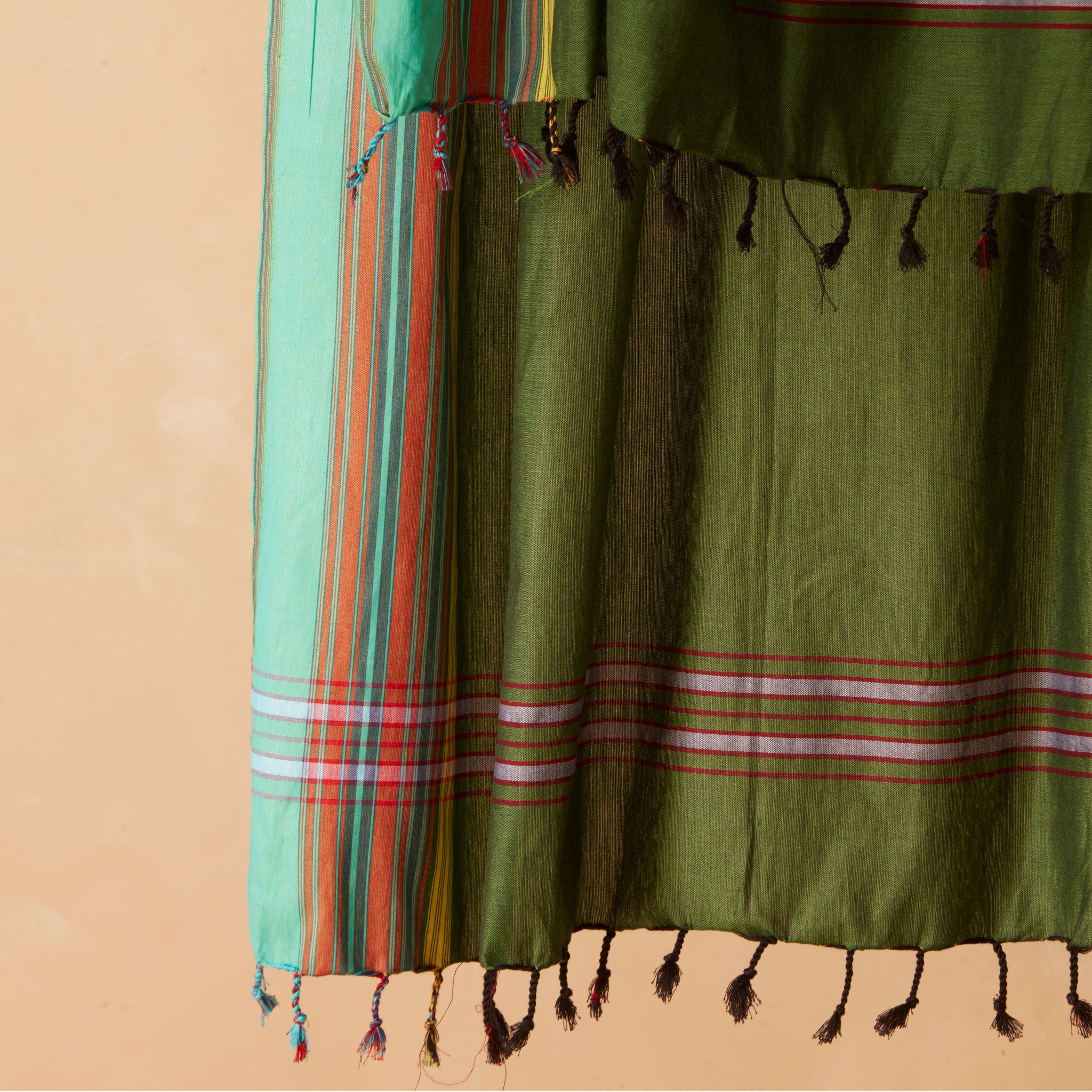 A close-up of an olive green sarong/pareo with horizontal striped borders in red, white, and pink, finished with dark tassels, shown against a warm beige background.