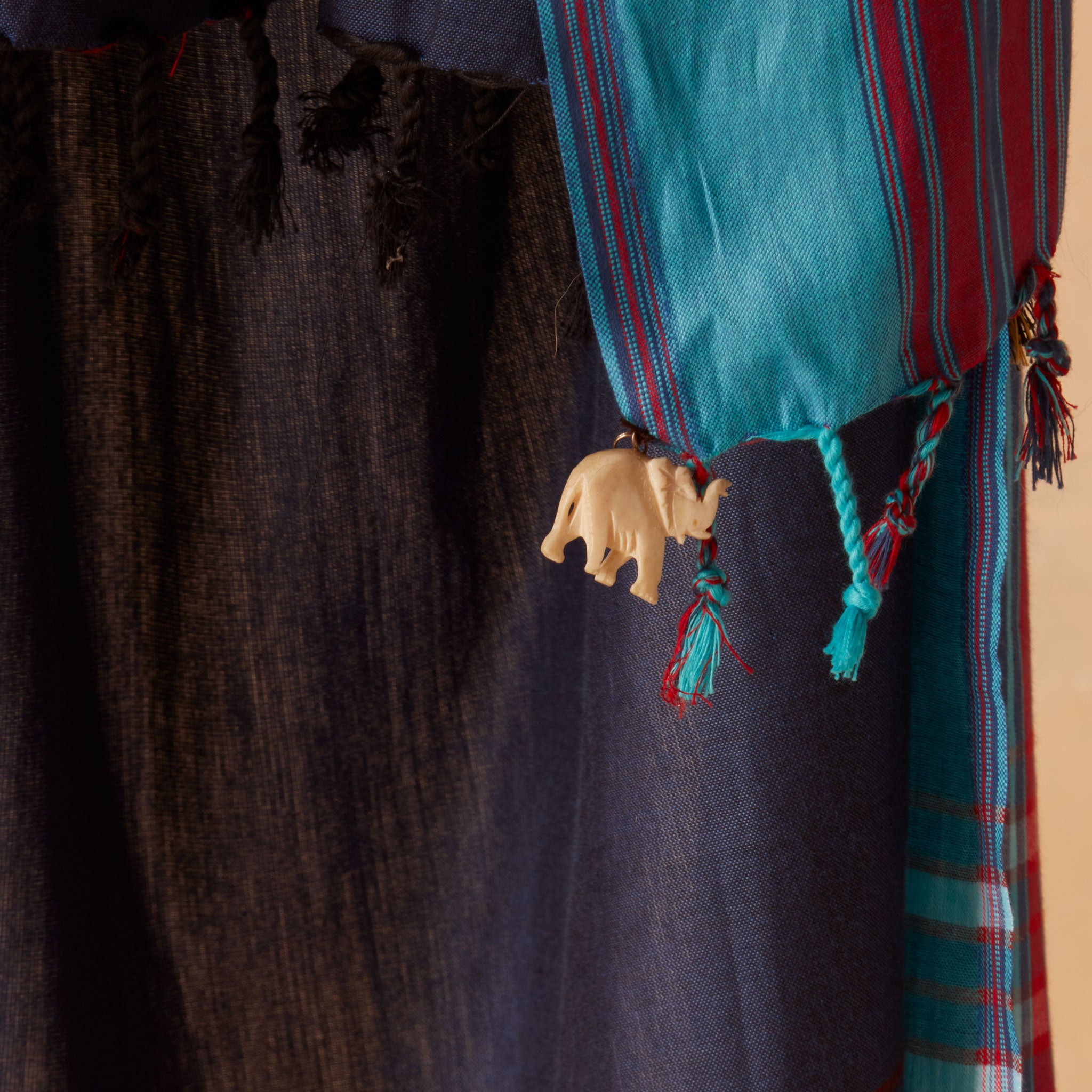 A close-up of a navy blue sarong/pareo with turquoise and red strips with braided tassels and a small elephant-shaped hanging charm.