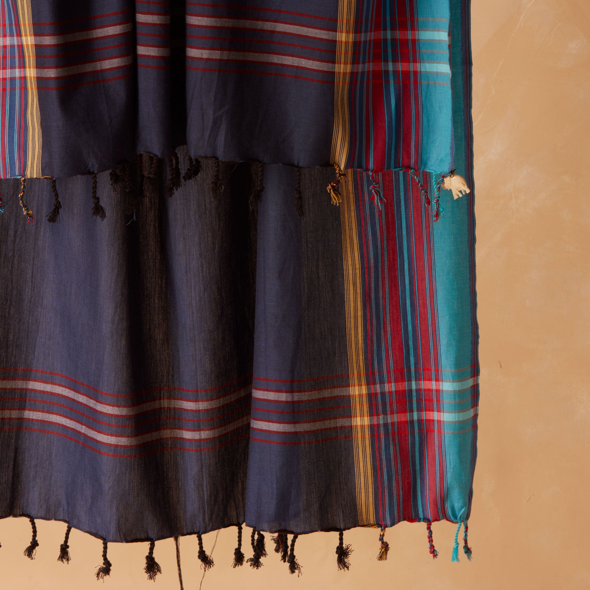 A dark navy blue woven sarong/pareo with vertical teal, red, yellow, and gold stripes and fringed tassels against a warm beige background.