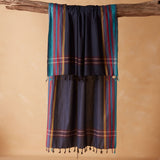 A dark navy blue woven sarong/pareo with vertical teal, red, yellow, and gold stripes and fringed tassels, draped over a rustic wooden branch against a warm beige background.