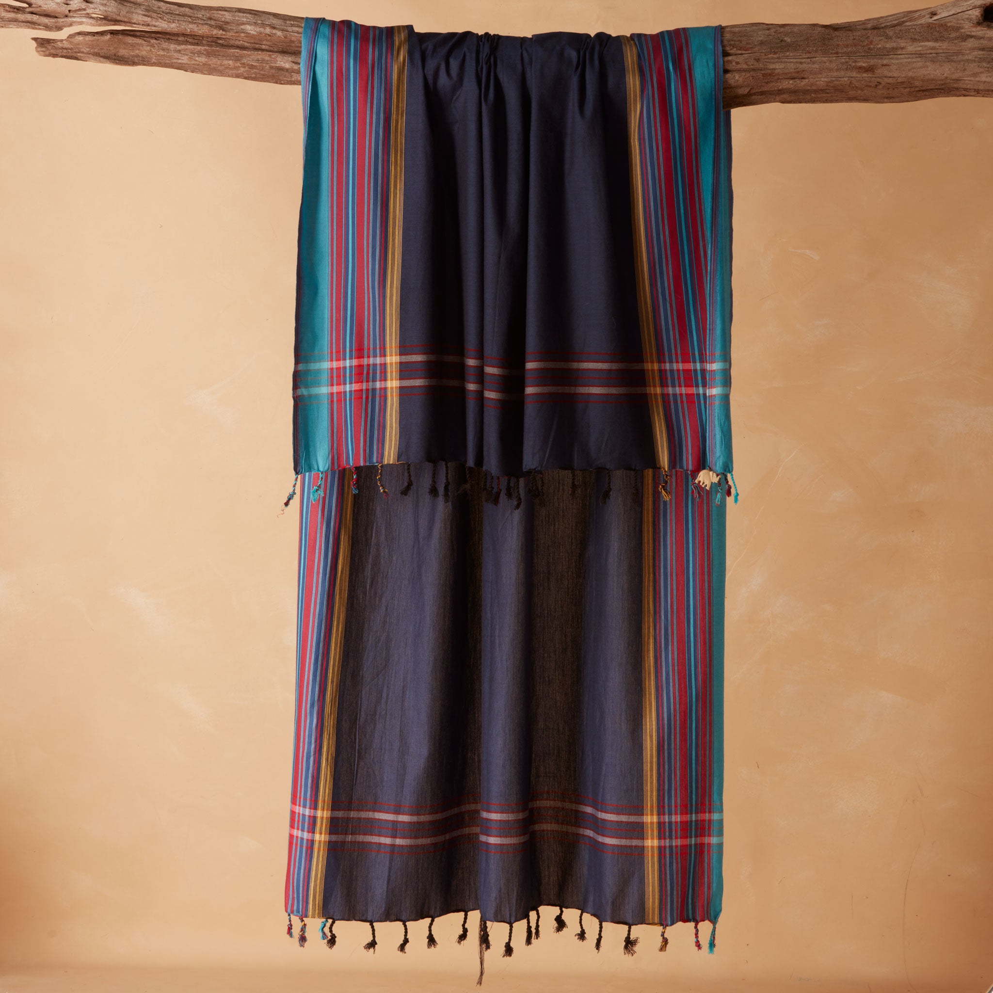 A dark navy blue woven sarong/pareo with vertical teal, red, yellow, and gold stripes and fringed tassels, draped over a rustic wooden branch against a warm beige background.
