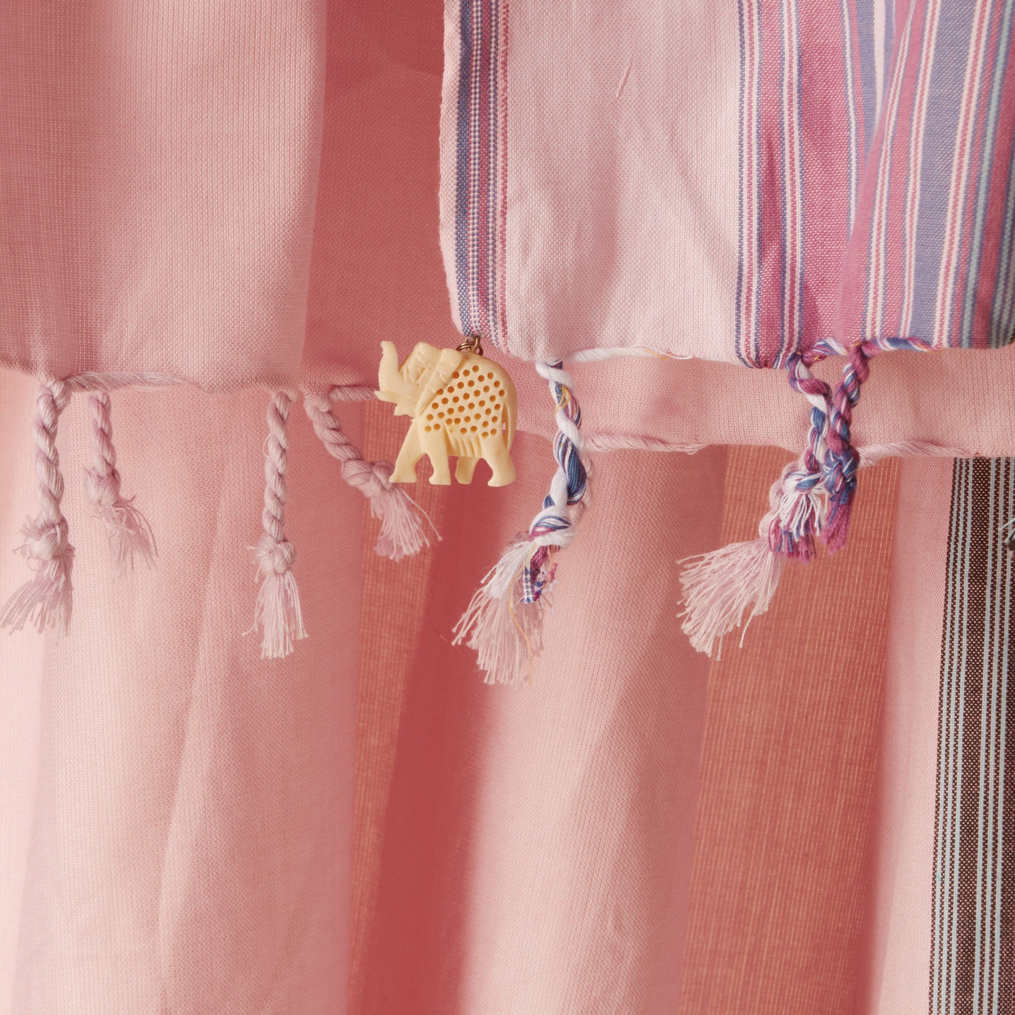 A close up of a cream colored carved elephant charm on the fringe of a pink woven pareo/sarong with vertical striped borders in purple, white, and blue.