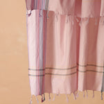 A pink woven pareo/sarong with vertical striped borders in purple, white, and blue with twisted tassel fringe against a warm beige background.