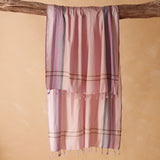 A pink woven pareo/sarong with vertical striped borders in purple, green, and blue, hanging from a rustic wooden branch against a warm beige background.