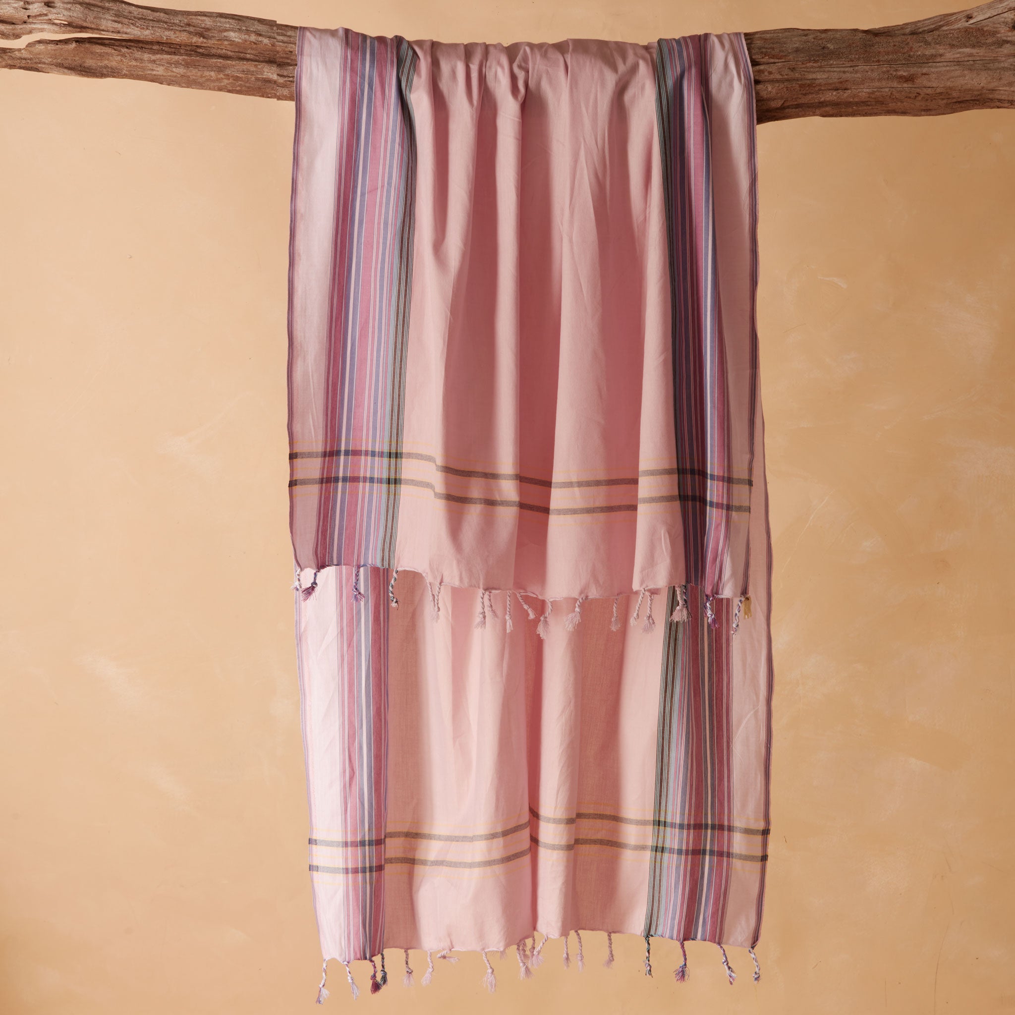 A pink woven pareo/sarong with vertical striped borders in purple, green, and blue, hanging from a rustic wooden branch against a warm beige background.