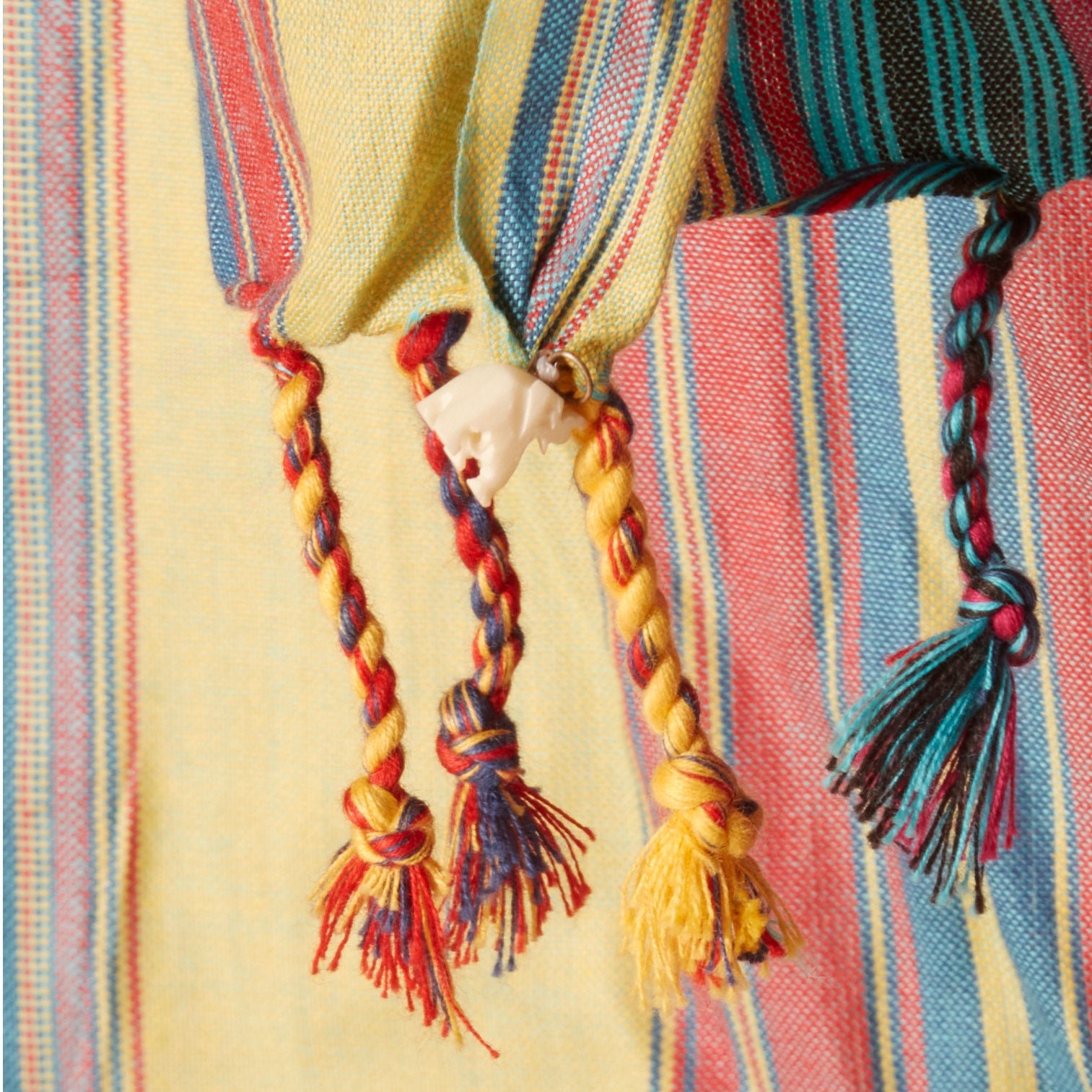 A pink pareo/sarong with vertical stripes in yellow, teal, and red with twisted tassel fringe and a cream colored carved elephant charm. 