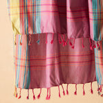 A close up of  pink pareo/sarong with vertical stripes in yellow, teal, and red with fringe against a warm beige background.