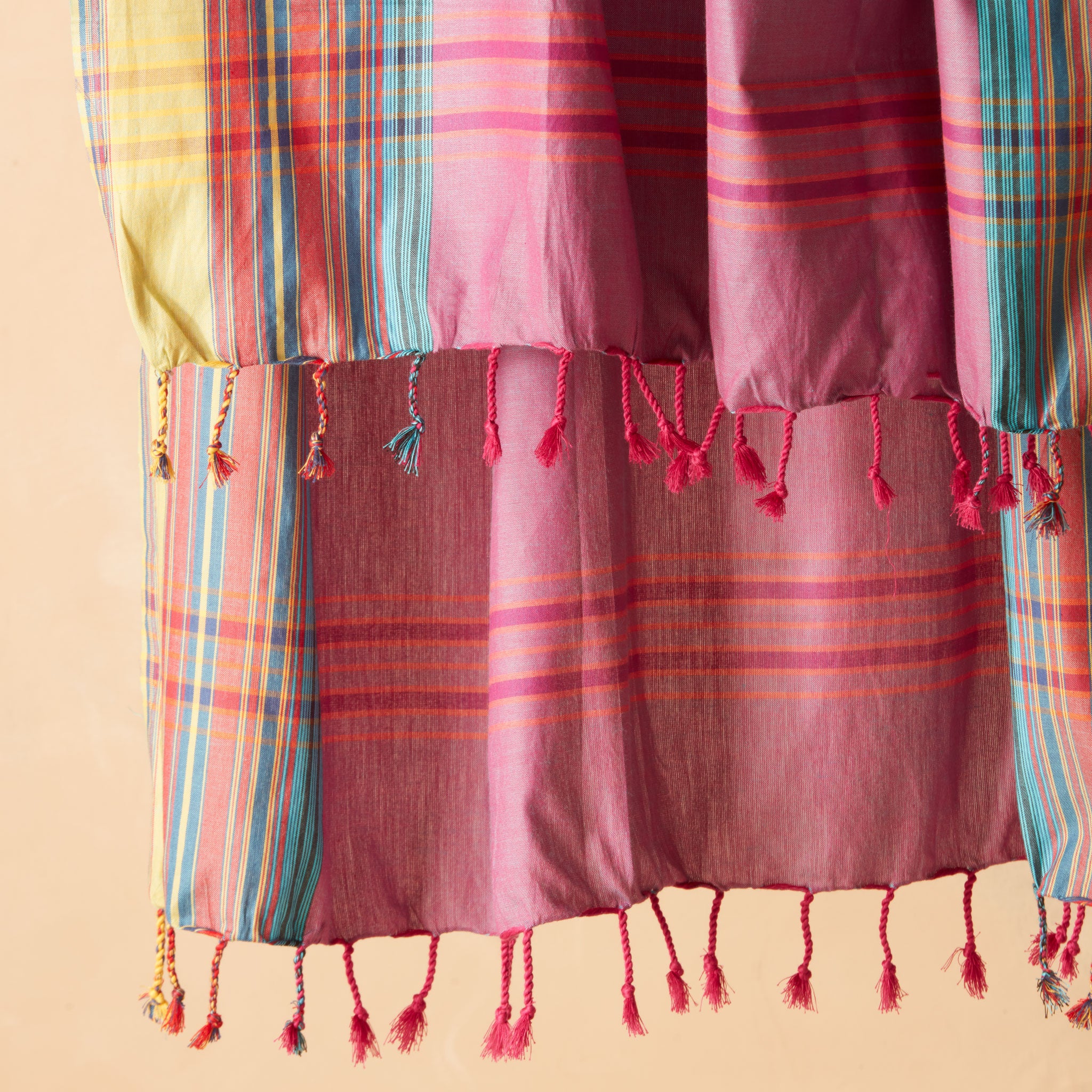 A close up of  pink pareo/sarong with vertical stripes in yellow, teal, and red with fringe against a warm beige background.
