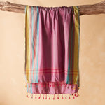 A pink pareo/sarong with vertical stripes in yellow, teal, and red, draped over a rustic wooden branch against a warm beige background.