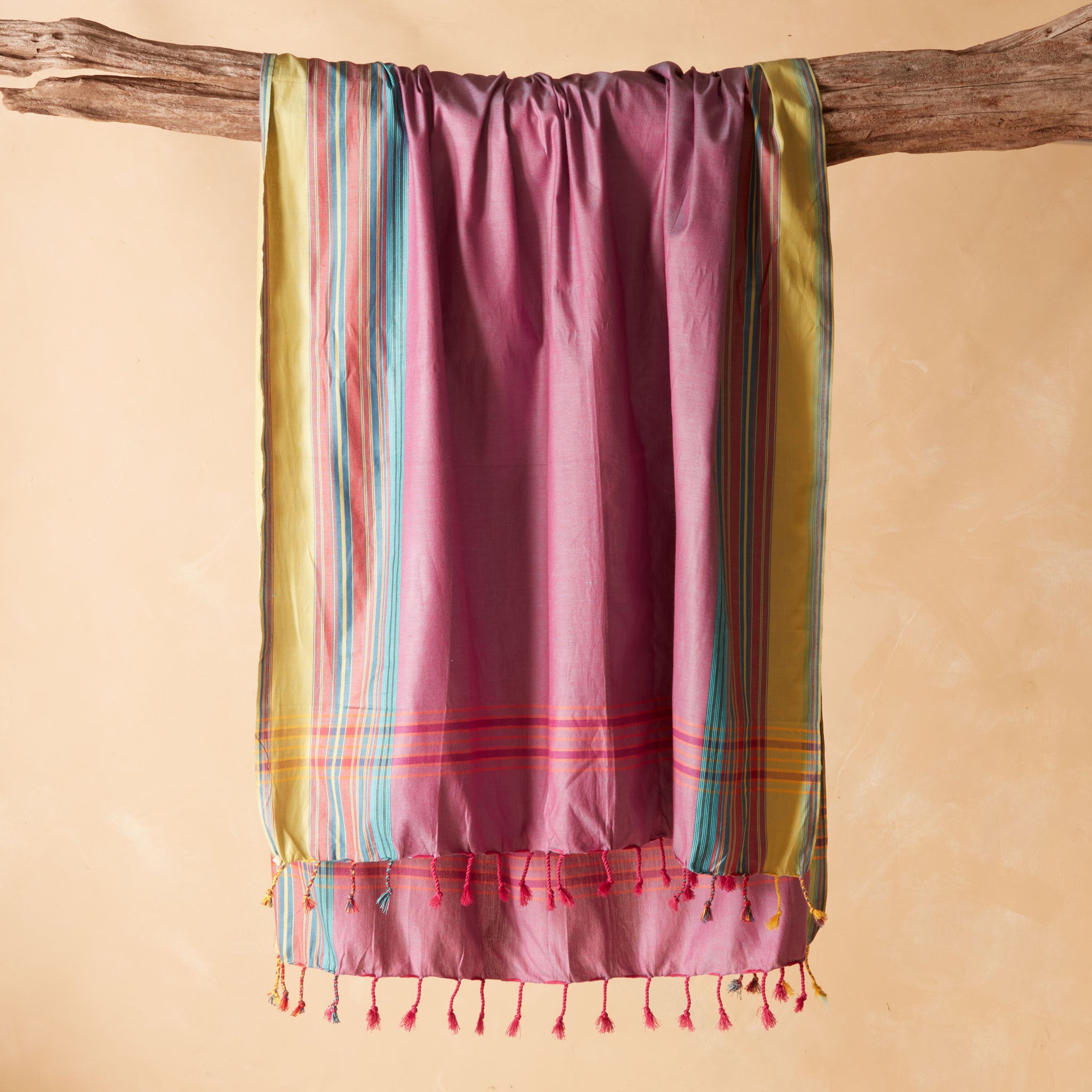 A pink pareo/sarong with vertical stripes in yellow, teal, and red, draped over a rustic wooden branch against a warm beige background.