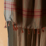 A close up of the fringe of a draped sarong/pareo in brown and grey with red striped bands and tasseled fringe, against a warm beige background. 