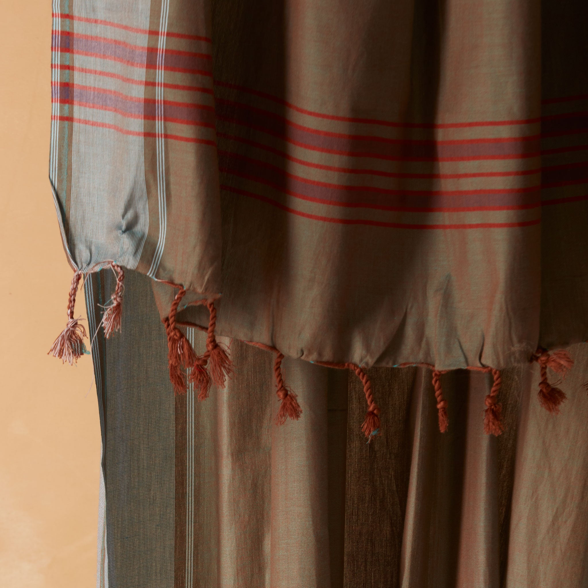 A close up of the fringe of a draped sarong/pareo in brown and grey with red striped bands and tasseled fringe, against a warm beige background. 