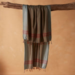 A draped sarong/pareo in brown and grey with red striped bands and tasseled fringe, hanging from a rustic wooden branch against a warm beige background. 