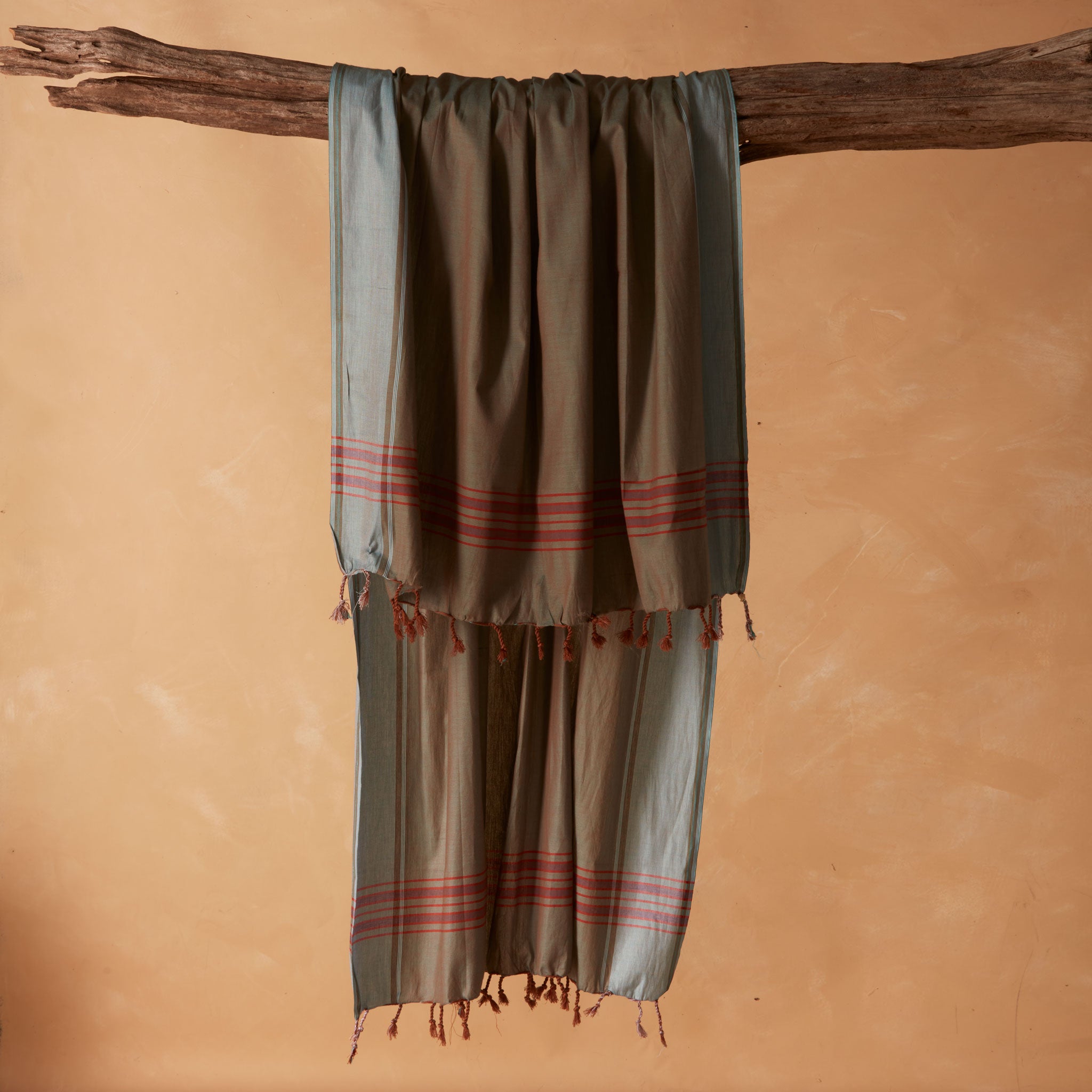 A draped sarong/pareo in brown and grey with red striped bands and tasseled fringe, hanging from a rustic wooden branch against a warm beige background. 
