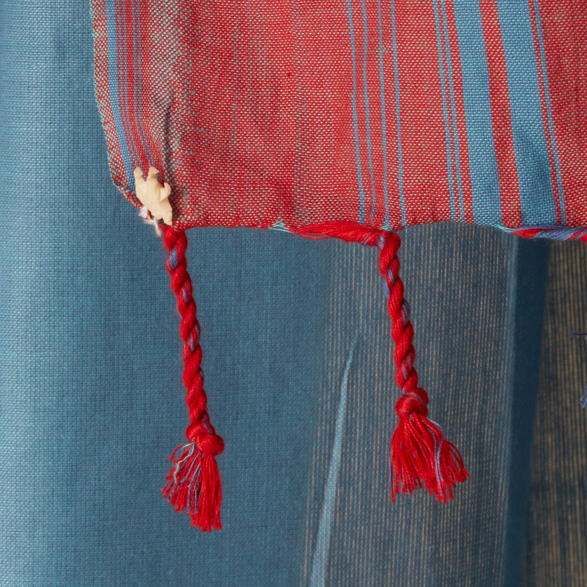 A close up of the fringe of the a teal blue and red striped/plaid patterned sarong/pareo with a cream colored elephant charm. 