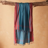 A teal blue and red striped/plaid patterned sarong/pareo with tassels hanging from a rustic wooden branch against a warm beige background.