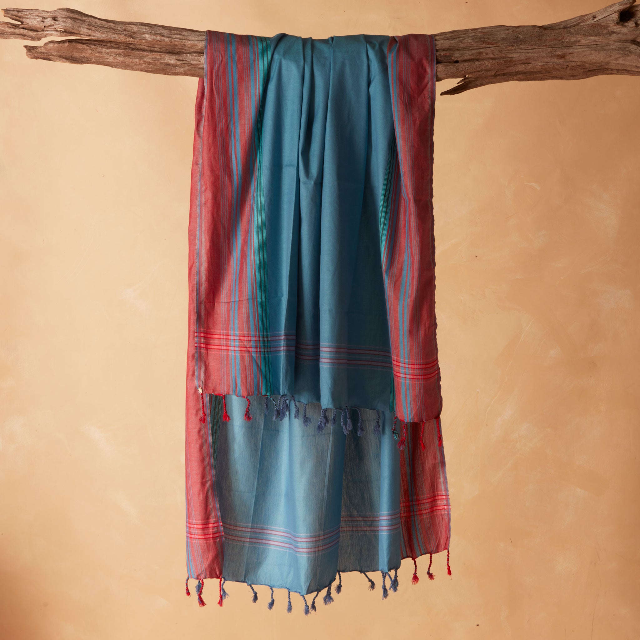 A teal blue and red striped/plaid patterned sarong/pareo with tassels hanging from a rustic wooden branch against a warm beige background.
