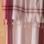A close up of a white, red and pink striped cotton pareo/sarong with fringed tassels against a warm beige background. 