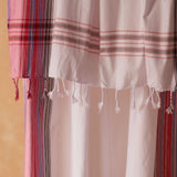 A close up of a white, red and pink striped cotton pareo/sarong with fringed tassels against a warm beige background. 
