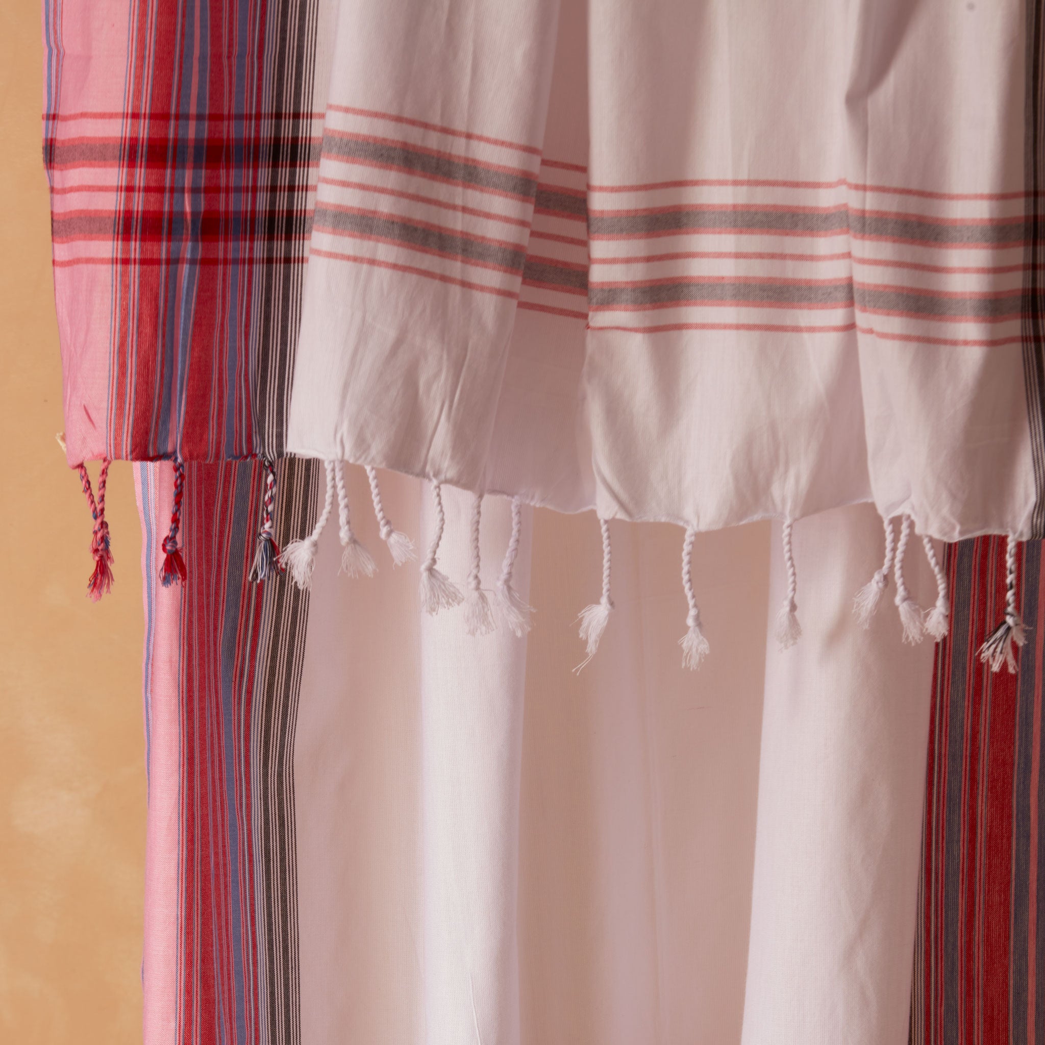 A close up of a white, red and pink striped cotton pareo/sarong with fringed tassels against a warm beige background. 