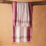A white and pink striped cotton pareo/sarong with fringed tassels hanging from a rustic wooden branch against a warm beige background.