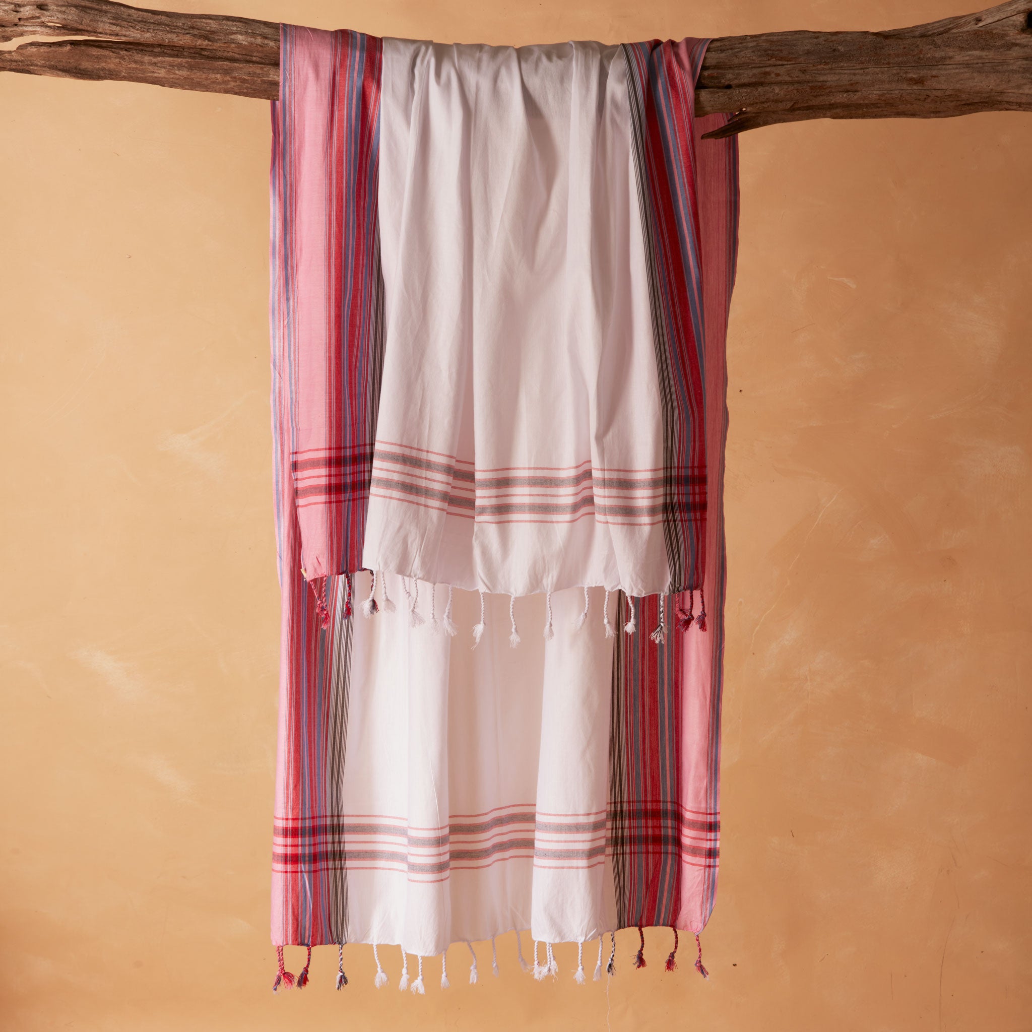 A white and pink striped cotton pareo/sarong with fringed tassels hanging from a rustic wooden branch against a warm beige background.