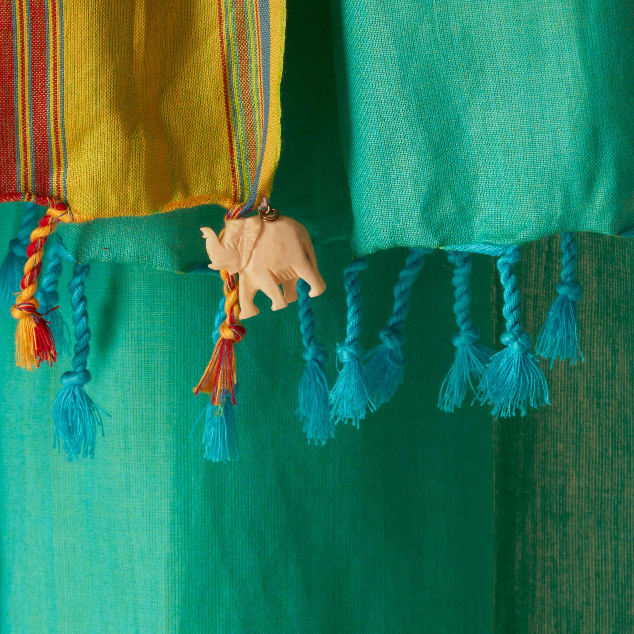 A close-up of colorful fabric tassels  of a bright turquoise green sarong/pareo, including a yellow striped section, a turquoise section, and a small carved elephant charm.