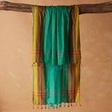 A woven pareo/sarong in bright turquoise green with yellow side panels and red striped accents, hanging from a rustic wooden branch against a warm beige background.