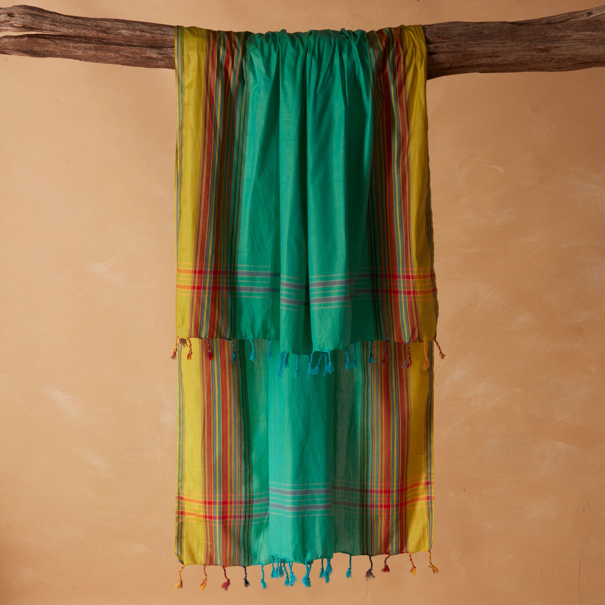 A woven pareo/sarong in bright turquoise green with yellow side panels and red striped accents, hanging from a rustic wooden branch against a warm beige background.