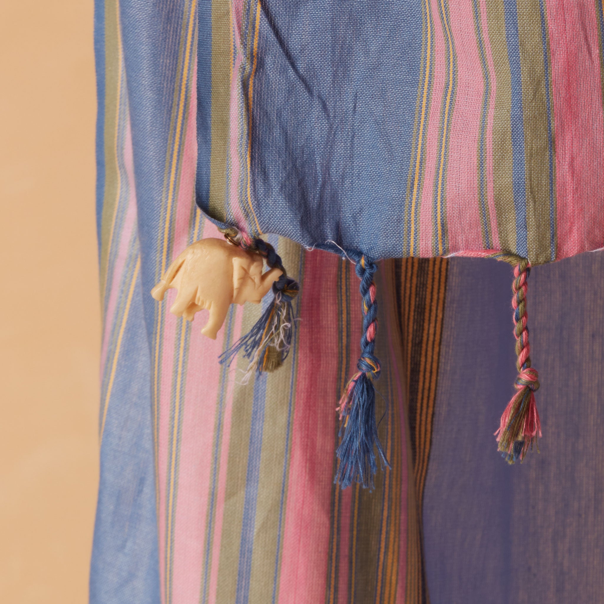 A close-up of a striped sarong/pareo in shades of blue, pink, and green, featuring braided tassels and a small elephant-shaped hanging charm.