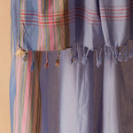 A close-up of a light blue and lavender sarong/pareo with pink red stripes, and green multicolor vertical plaid sections, and braided tassels.