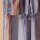 A close-up of a light blue and lavender sarong/pareo with pink red stripes, and green multicolor vertical plaid sections, and braided tassels.