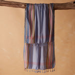 A blue striped cotton sarong/pareo with multi colored stripes with tassels hanging from a rustic wooden branch against a warm beige background.
