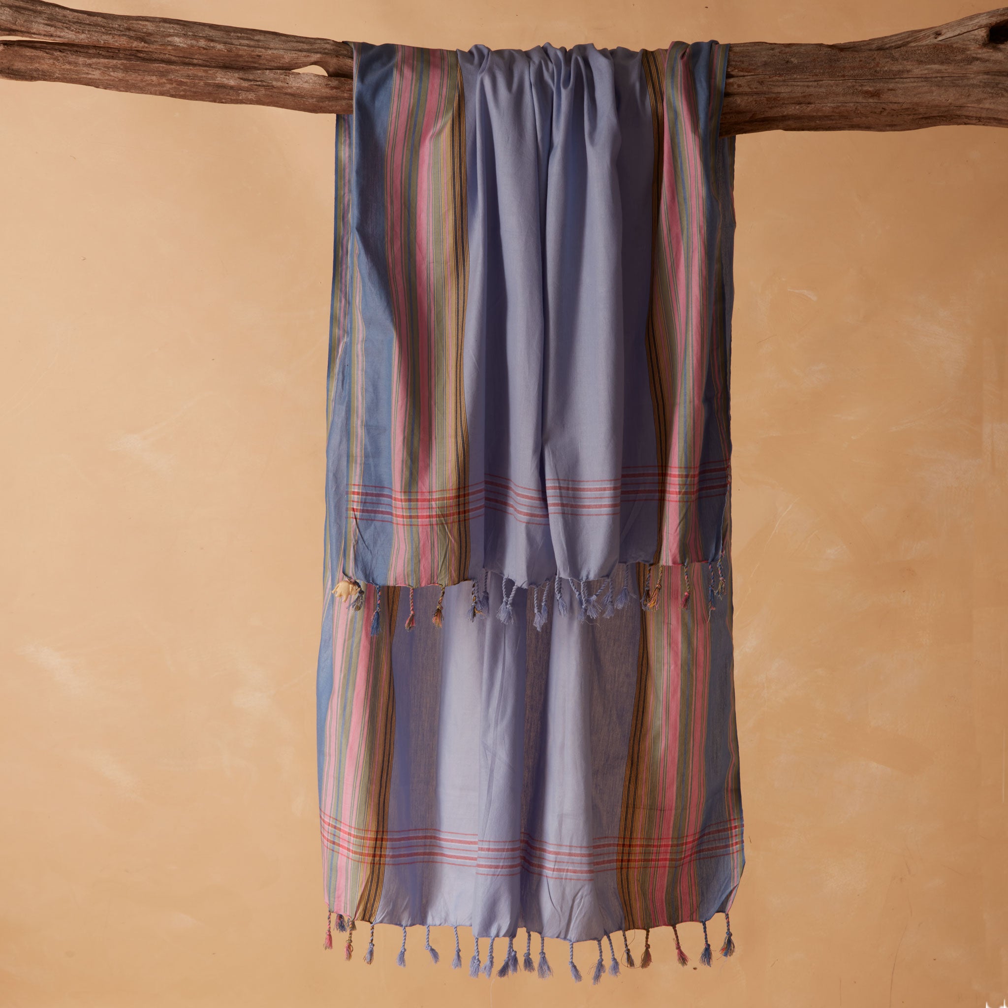 A blue striped cotton sarong/pareo with multi colored stripes with tassels hanging from a rustic wooden branch against a warm beige background.