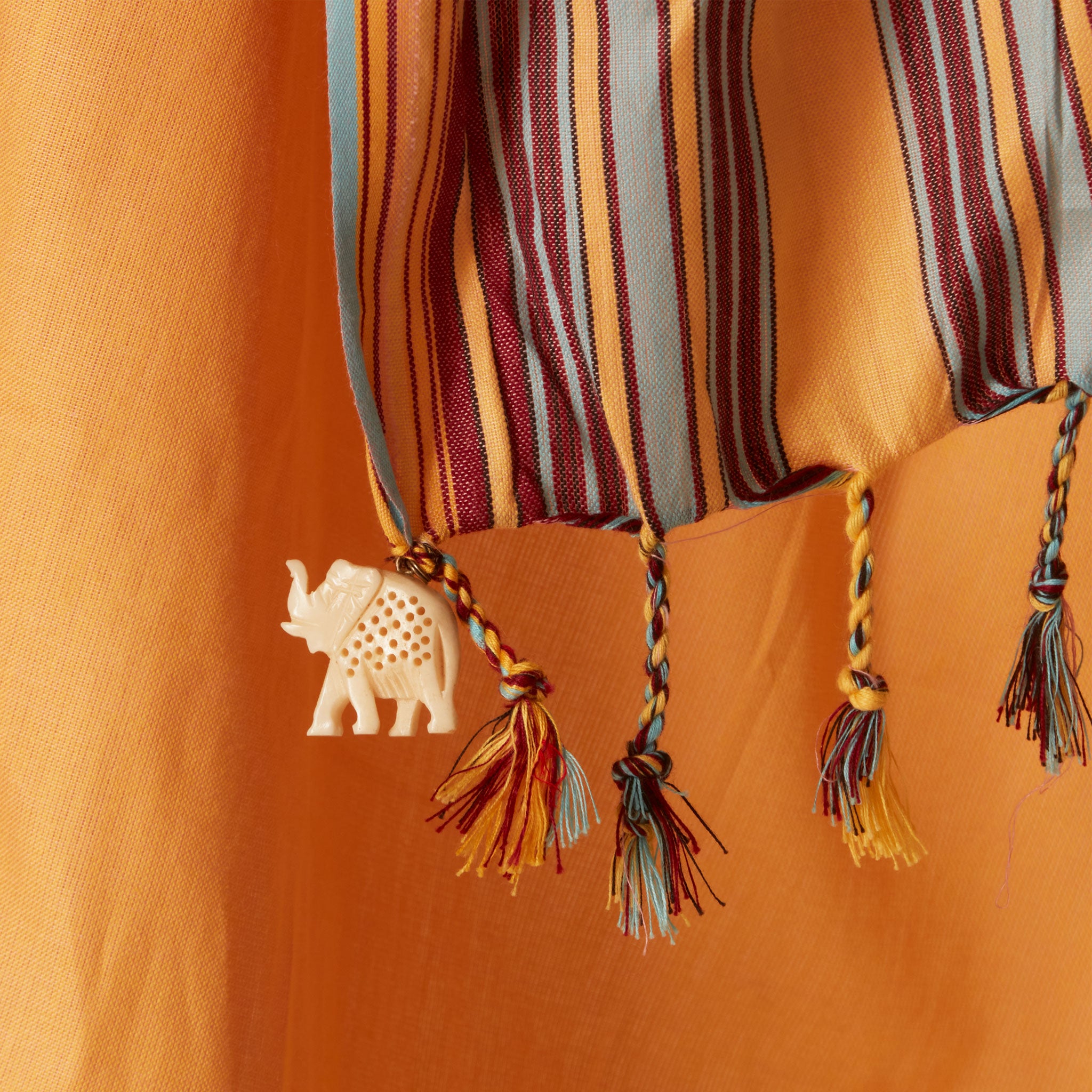 A close up of twisted fringe and a cream colored elephant charm  of a A yellow orange cotton pareo/sarong with multicolored striped borders.