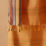 A close-up of an orange sarong/pareo with multicolor stripes and tassels, draped against a warm beige background.